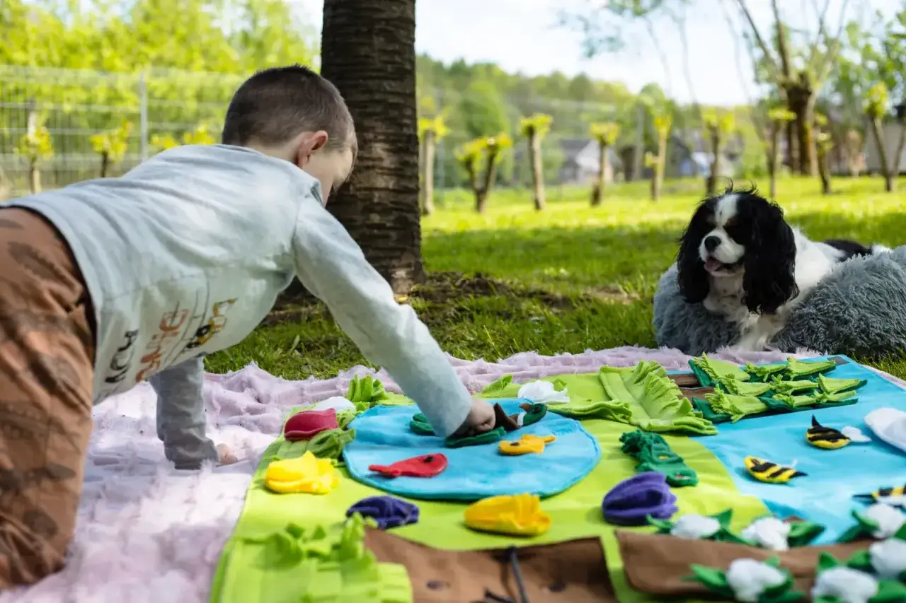 Chłopiec bawi się na macie edukacyjnej, a obok niego leży pies terapeutyczny, obserwując go z uwagą.